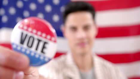 Portrait-of-biracial-man-in-front-of-american-flag-holding-badge-with-vote-text,-slow-motion
