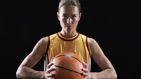 Young-Caucasian-woman-poses-confidently-in-a-basketball-uniform,-holding-a-ball-on-a-black-backgroun