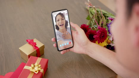 Caucasian-woman-holding-holding-smartphone-with-caucasian-woman-on-screen,-gifts-on-desk
