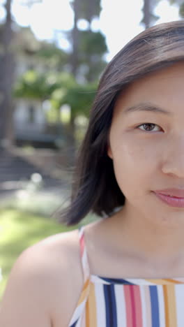 Half-face-of-happy-asian-woman-with-dark-hair-standing-and-smiling-in-sunny-garden,-slow-motion