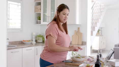 Happy-plus-size-biracial-woman-making-meal-with-vegetables-in-kitchen-and-using-tablet,-slow-motion