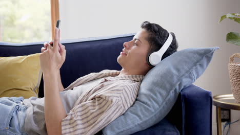 Happy-biracial-man-in-headphones-lying-on-couch-using-smartphone,-slow-motion