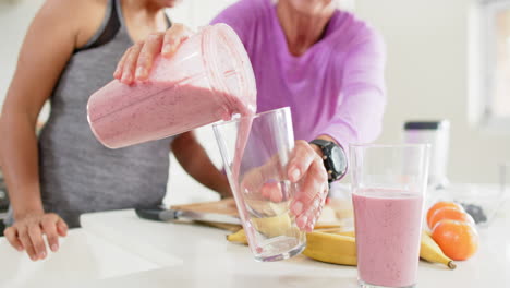 Two-happy-diverse-senior-women-pouring-cocktail-into-glasses-in-kitchen,-slow-motion