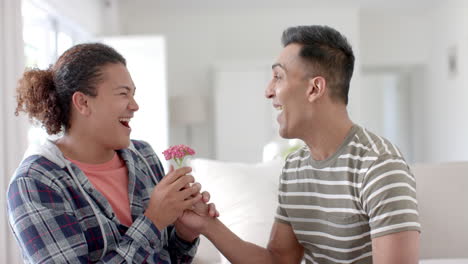 Happy-diverse-gay-male-couple-sitting-on-couch-and-giving-flower-in-living-room,-slow-motion
