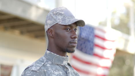 Ernster-Afroamerikanischer-Soldat,-Der-An-Einem-Sonnigen-Tag-Mit-Der-Flagge-Im-Hintergrund-Steht,-Zeitlupe
