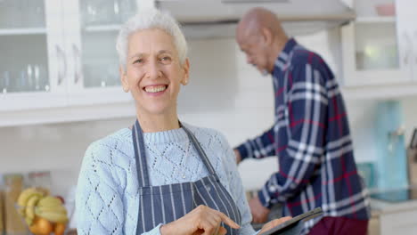Portrait-of-happy-senior-biracial-couple-cooking-and-using-tablet-in-kitchen,-slow-motion