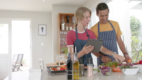 Happy-diverse-couple-preparing-fresh-vegetables-and-using-tablet-in-kitchen,-slow-motion