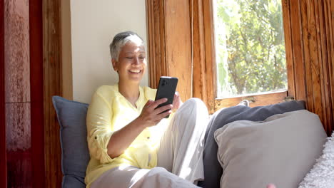Glückliche-ältere-Frau-Mit-Gemischter-Abstammung,-Die-Auf-Der-Couch-Sitzt-Und-Zu-Hause-Ihr-Smartphone-Benutzt,-Zeitlupe