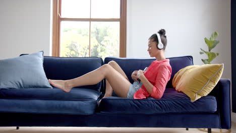 Happy-biracial-teenage-girl-in-headphones-relaxing-on-couch-with-smartphone,-copy-space,-slow-motion