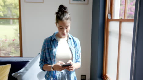 Happy-biracial-teenage-girl-standing-in-sunny-room-using-smartphone,-copy-space,-slow-motion