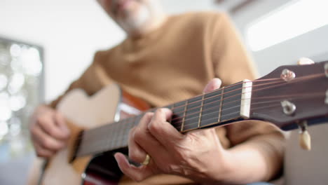 Midsection-of-senior-biracial-man-sitting-playing-guitar-at-home,-selective-focus,-slow-motion