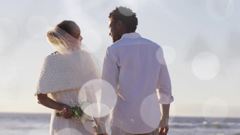 Animation-of-light-spots-over-african-american-bride-and-groom-holding-hands-on-beach-at-wedding