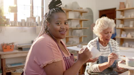 Two-diverse-female-potters-glazing-clay-jug-and-smiling-in-pottery-studio,-slow-motion