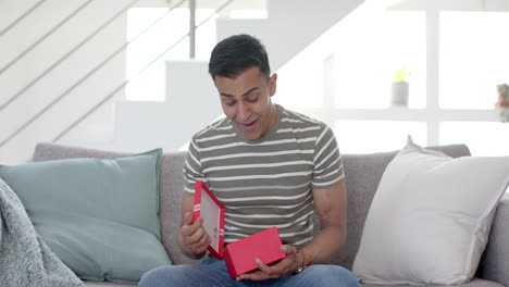 Happy-surprised-biracial-man-having-video-call-and-opening-present-in-sunny-living-room,-slow-motion