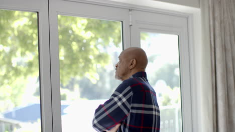 Thoughtful-senior-biracial-man-looking-through-window-at-home,-slow-motion