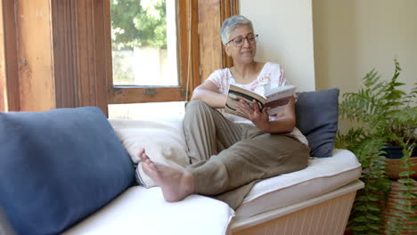 Glückliche-ältere-Frau-Mit-Gemischter-Abstammung-Auf-Der-Couch,-Die-Zu-Hause-Am-Fenster-Ein-Buch-Liest,-Zeitlupe
