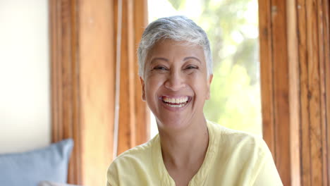 Portrait-of-happy-senior-biracial-woman-sitting-on-couch-at-window-at-home,-slow-motion