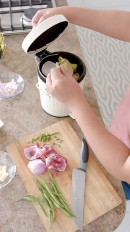 Vertical-video-of-plus-size-biracial-woman-composting-vegetable-waste-in-kitchen,-slow-motion