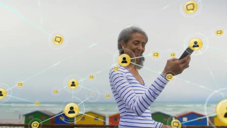 Animation-of-network-of-connections-with-icons-over-biracial-woman-using-smartphone-by-sea