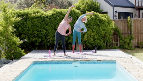 Two-happy-diverse-senior-women-stretching-and-smiling-by-pool-in-sunny-garden,-slow-motion