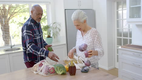 Happy-senior-biracial-couple-unpacking-shopping-bags-in-kitchen,-slow-motion