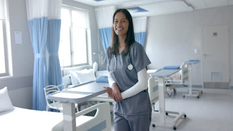 Portrait-of-happy-biracial-female-doctor-in-scrubs-smiling-in-hospital-ward,-copy-space,-slow-motion