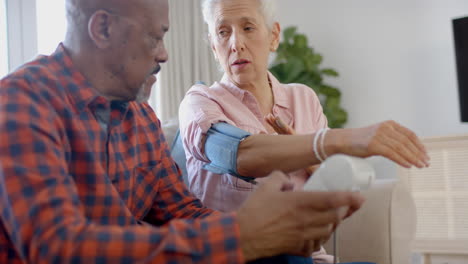 Ernsthaftes-älteres-Biracial-Paar,-Das-Auf-Der-Couch-Sitzt-Und-Zu-Hause-Den-Blutdruck-überprüft,-Zeitlupe