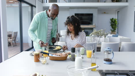 Happy-biracial-couple-having-breakfast-with-scrambled-eggs-in-kitchen,-slow-motion