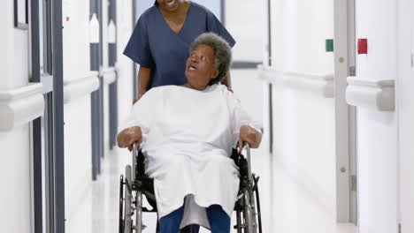 African-american-female-doctor-walking-with-senior-woman-in-wheelchair-in-hospital,-slow-motion