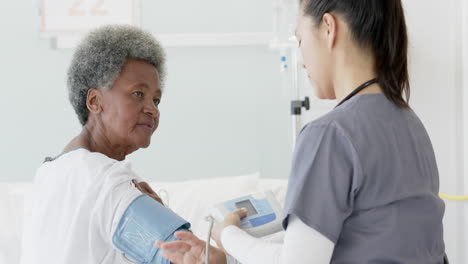 Diverse-female-doctor-testing-blood-pressure-of-senior-female-patient-in-hospital-room,-slow-motion