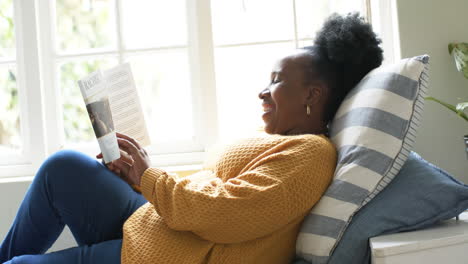 Glückliche-Afroamerikanische-Seniorin,-Die-Im-Sonnigen-Zimmer-Liegt,-Ein-Buch-Liest-Und-Lächelt,-Zeitlupe