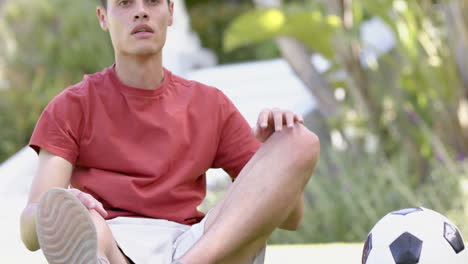 Focused-biracial-man-sitting-with-football,-touching-toes-in-sunny-garden,-copy-space,-slow-motion