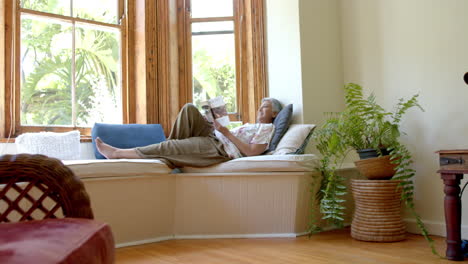 Glückliche-ältere-Frau-Mit-Gemischter-Abstammung-Auf-Der-Couch,-Die-Zu-Hause-Am-Fenster-Ein-Buch-Liest,-Zeitlupe