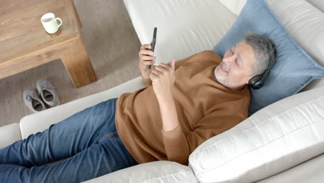 Glücklicher-älterer-Mann-Mit-Gemischter-Abstammung-Auf-Der-Couch-Mit-Kopfhörern,-Der-Musik-Auf-Dem-Smartphone-Hört,-Zeitlupe