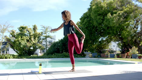 Happy-biracial-woman-practicing-yoga-by-pool,-holding-foot-in-sunny-garden,-copy-space,-slow-motion
