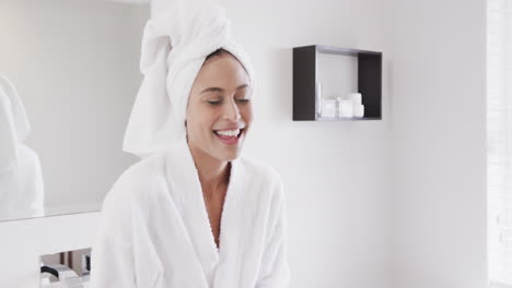Portrait-of-happy-biracial-woman-wearing-towel-on-head-using-smartphone-in-bathroom,-slow-motion
