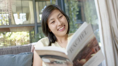 Feliz-Mujer-Asiática-Sentada-En-El-Sofá-Y-Leyendo-Un-Libro-En-Una-Soleada-Sala-De-Estar,-Cámara-Lenta