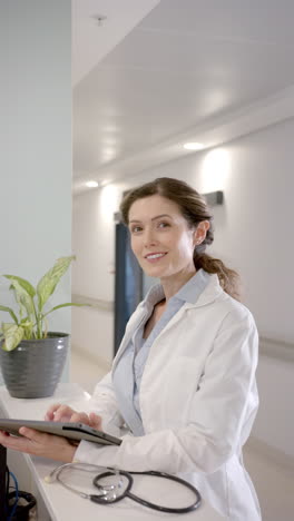 Vertical-video-portrait-of-happy-caucasian-female-doctor-using-tablet-at-reception-desk,-slow-motion