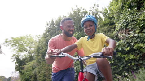 Glücklicher-Afroamerikanischer-Vater-Bringt-Sohn-Das-Fahrradfahren-Im-Sonnigen-Garten-Bei,-Zeitlupe