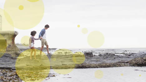 Animation-of-light-spots-over-biracial-couple-holding-hands-and-walking-on-beach