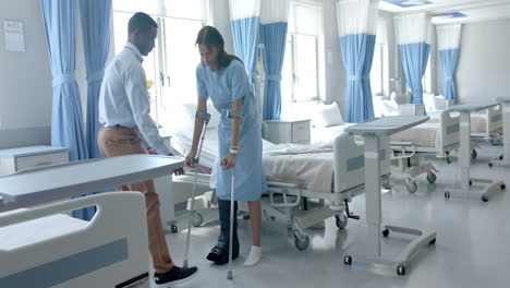 Diverse-male-doctor-helping-happy-female-patient-walk-with-crutches-in-ward,-copy-space,-slow-motion