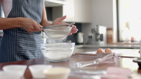Happy-biracial-woman-biracial-woman-in-apron-sieving-flour,-baking-in-kitchen,-slow-motion