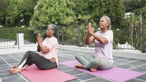 Senior-african-american-female-friends-practicing-yoga-meditation-sitting-on-balcony,-slow-motion