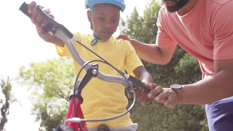 Padre-Afroamericano-Enseñando-A-Su-Hijo-A-Andar-En-Bicicleta-En-Un-Jardín-Soleado,-Cámara-Lenta