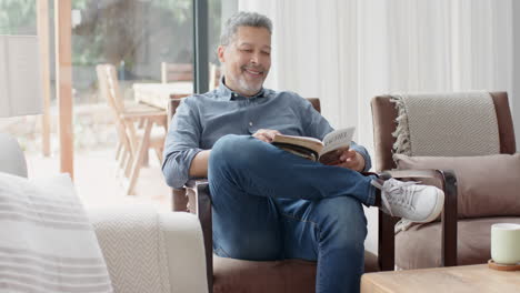 Happy-senior-biracial-man-sitting-in-armchair-reading-book-in-living-room,-slow-motion