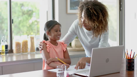 Happy-biracial-mother-and-daughter-using-laptop-for-schoolwork-in-kitchen,-slow-motion