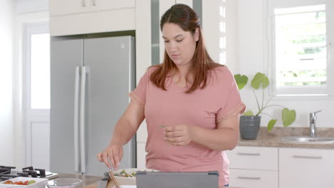 Happy-plus-size-biracial-woman-making-vegetable-salad-in-kitchen-and-using-tablet,-slow-motion