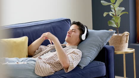 Happy-biracial-man-in-headphones-lying-on-couch-using-smartphone,-copy-space,-slow-motion