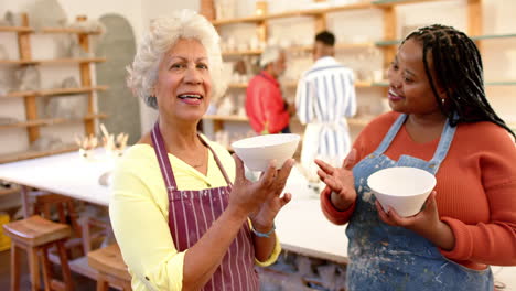 Happy-diverse-female-potters-holding-bowls-and-smiling-in-pottery-studio,-slow-motion