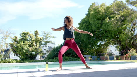 Happy-biracial-woman-practicing-yoga-standing-by-pool-in-sunny-garden,-copy-space,-slow-motion